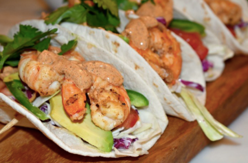 Three shrimp tacos topped with sauce, avocado, tomatoes, and cilantro on a wooden platter.