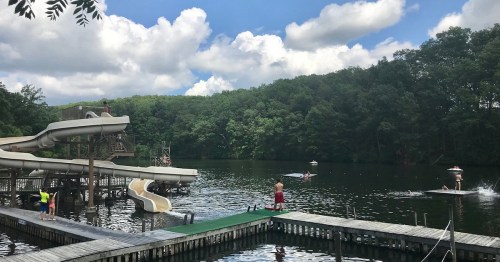 A scenic lake with water slides, people swimming, and boats, surrounded by lush green trees and a blue sky.