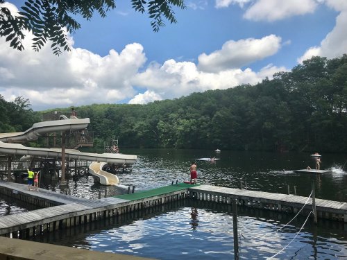 A scenic lake with water slides, people swimming, and boats, surrounded by lush green trees and a blue sky with clouds.