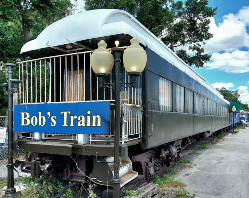 A vintage train car named "Bob's Train" with a blue sign, parked beside a fence under a partly cloudy sky.