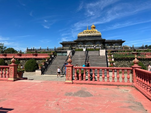 A grand building with a golden dome, surrounded by lush greenery and steps, under a clear blue sky.