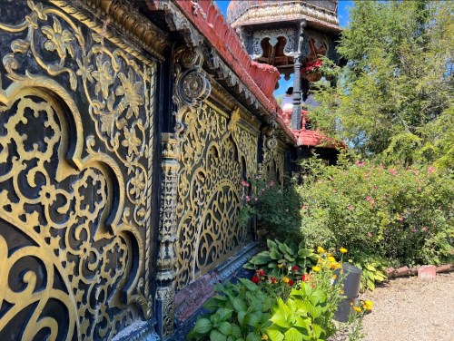 Intricate gold and black carvings on a wall, surrounded by vibrant flowers and greenery under a clear blue sky.