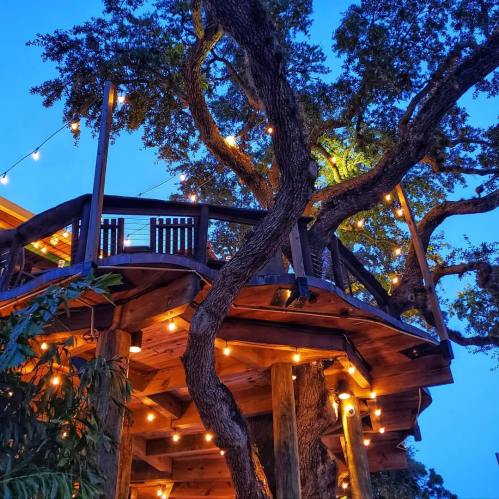 A beautifully lit treehouse nestled in a large tree, surrounded by greenery and evening sky.