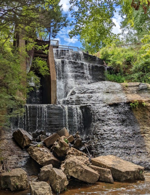 A cascading waterfall flows over rocky terrain, surrounded by lush greenery and a blue sky with scattered clouds.