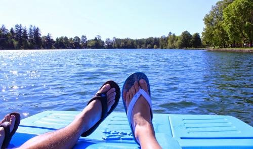 Two pairs of feet in flip-flops resting on a blue dock, overlooking a calm lake surrounded by trees on a sunny day.