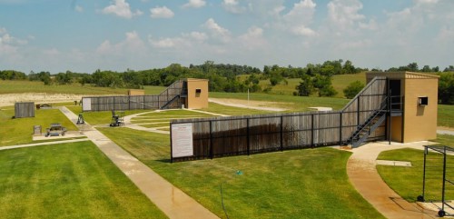 A wide view of a shooting range with two elevated platforms, grassy areas, and a clear blue sky in the background.