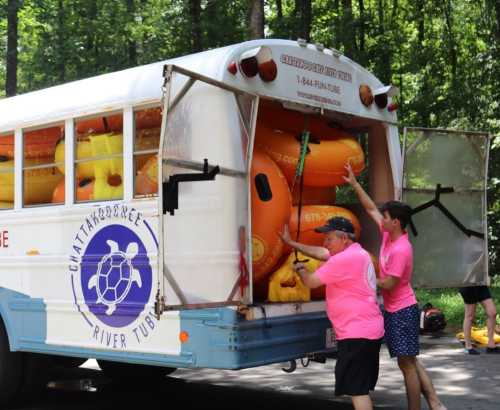 Two people in pink shirts load orange river tubes into the back of a bus in a wooded area.