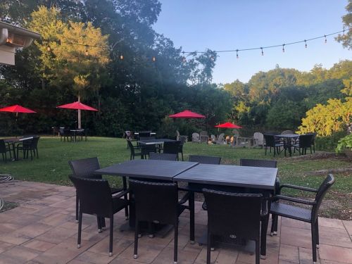Outdoor seating area with black tables and chairs, red umbrellas, and a green lawn surrounded by trees.