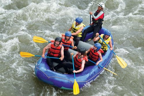 A group of six people in helmets and life jackets paddling a blue raft through turbulent water.