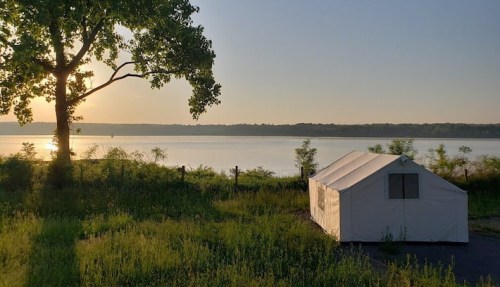 A white tent sits by a calm lake at sunset, surrounded by lush greenery and a large tree.