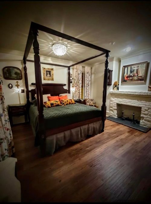 Cozy bedroom featuring a four-poster bed, decorative pillows, artwork, and a stone fireplace. Warm, inviting atmosphere.