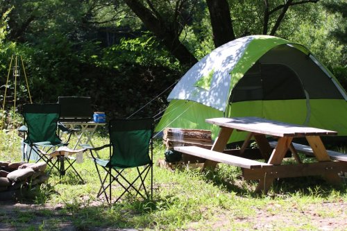 A green and gray tent set up in a grassy campsite with folding chairs and a picnic table nearby.