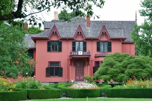 A large, red Victorian-style house with intricate architectural details, surrounded by lush greenery and colorful flowers.