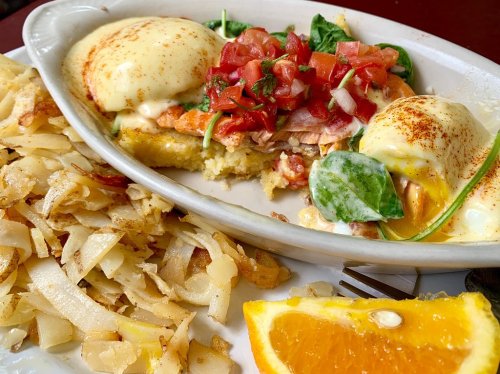 A plate of eggs Benedict with spinach, tomatoes, and hollandaise sauce, served with hash browns and an orange slice.