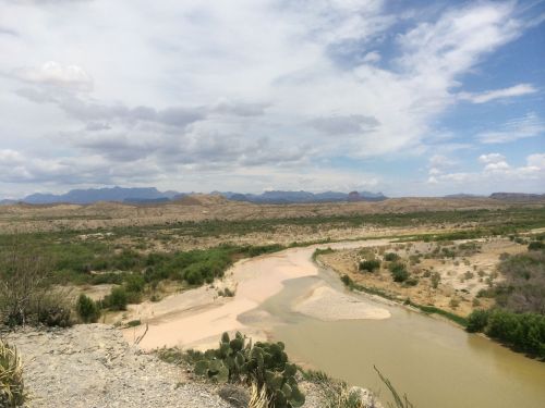 A scenic view of a river winding through arid land, with mountains in the background and scattered vegetation.
