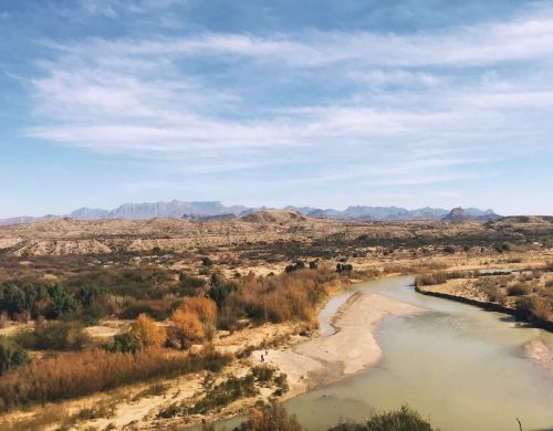 A scenic view of a winding river surrounded by arid land and distant mountains under a blue sky with wispy clouds.