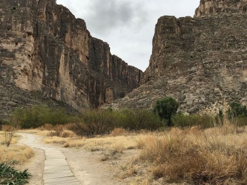 A winding path through a dry landscape, flanked by towering rocky cliffs and sparse vegetation under a cloudy sky.
