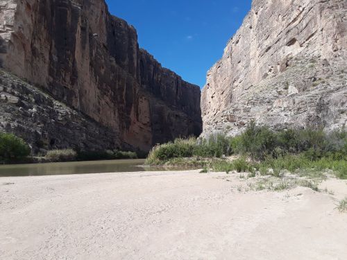 A sandy riverbank surrounded by towering rock cliffs under a clear blue sky.