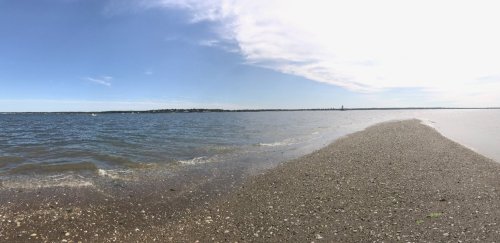A panoramic view of a calm shoreline with water, sand, and a clear blue sky in the background.