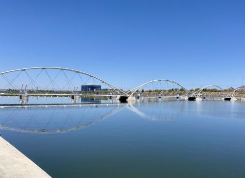 A modern bridge arches over a calm body of water, reflecting the clear blue sky and surrounding landscape.
