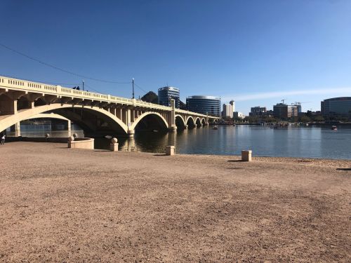 A bridge spans a calm body of water, with modern buildings and a clear blue sky in the background.