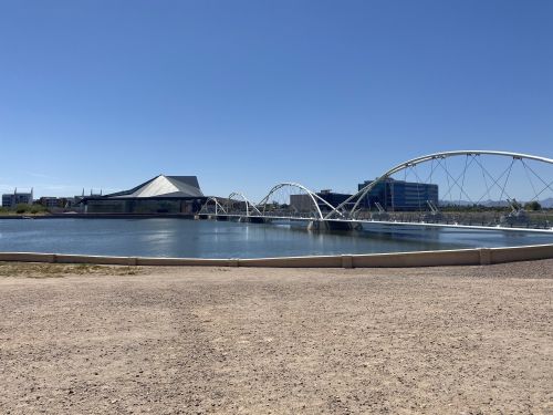 A modern bridge spans a calm body of water, with a contemporary building and clear blue sky in the background.