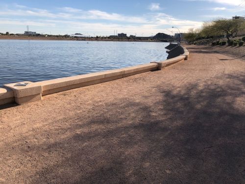 A sandy path along a calm lake, with buildings in the background and a clear blue sky.