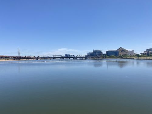 A calm river reflects a clear blue sky, with a bridge and modern buildings visible along the shoreline.