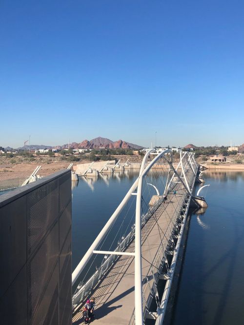 A modern bridge spans a calm river, with mountains in the background under a clear blue sky.