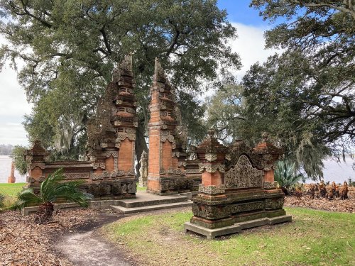 Balinese-style temple ruins surrounded by lush greenery and trees, near a body of water under a blue sky.