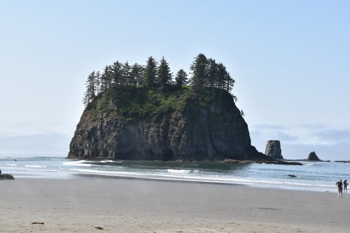 A rocky island with trees stands in the ocean, surrounded by sandy beach and gentle waves under a clear blue sky.