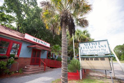 Exterior of a seafood restaurant with a sign reading "Stop Here for Good Food" and palm trees in the foreground.