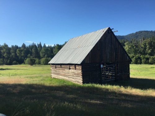 A rustic wooden barn with a metal roof stands in a grassy field, surrounded by trees and mountains under a clear blue sky.