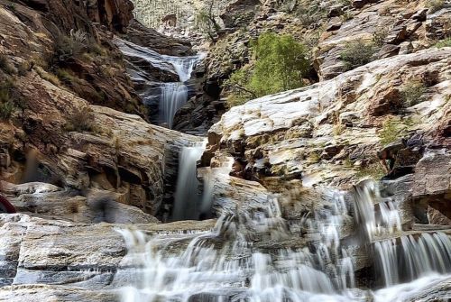 A cascading waterfall flows over rocky cliffs, surrounded by lush greenery and rugged terrain.