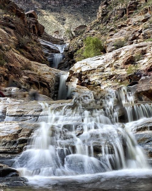 A cascading waterfall flows over rocky terrain, surrounded by lush greenery and steep cliffs.