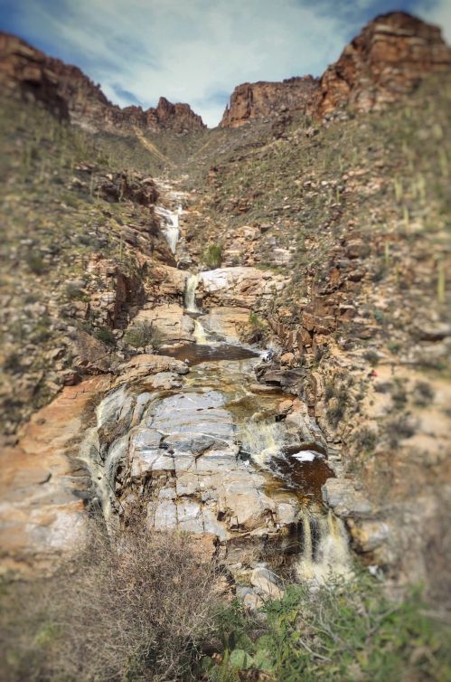 A scenic view of a rocky canyon with a waterfall cascading down, surrounded by desert vegetation and mountains.