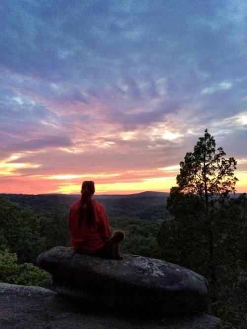 A person sits on a rock, gazing at a colorful sunset over a lush landscape.