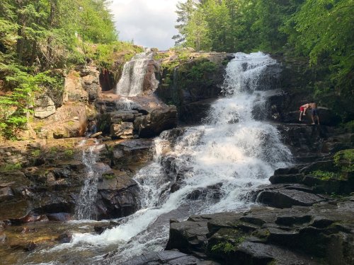 A scenic waterfall cascades over rocky terrain, surrounded by lush green trees and a person exploring nearby.