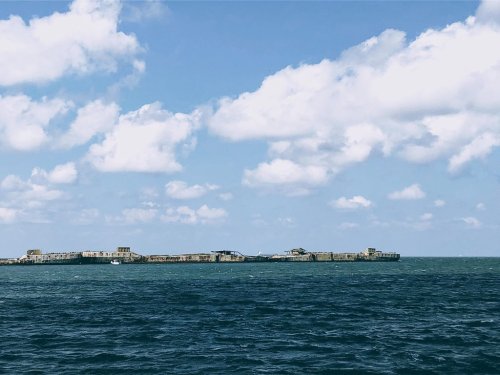 A distant view of a weathered structure on the water, surrounded by blue waves and a partly cloudy sky.