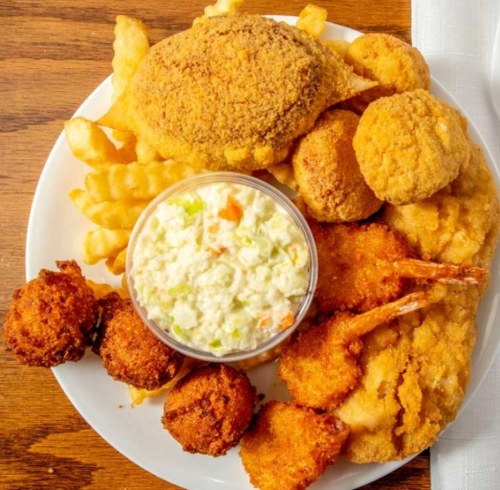 A plate of fried seafood, including fish, shrimp, hushpuppies, coleslaw, and crinkle-cut fries.