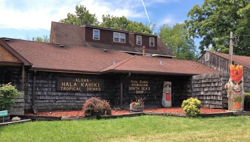 A wooden building with signs for "Hala Kahiki Tropical Drinks" and "Hawaiian South Seas Shop," featuring a tiki statue.