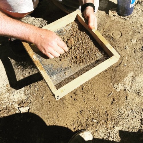A person sifts soil through a wooden screen, separating finer particles from larger clumps on the ground.