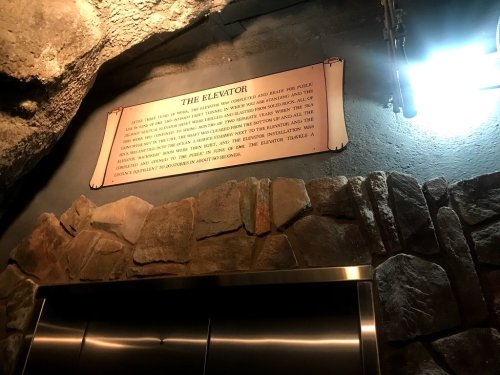 A stone wall with a sign about an elevator, illuminated by a light, in a cave-like setting.