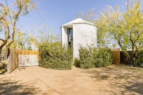 A modern cylindrical building surrounded by greenery and wooden fencing, set in a gravel landscape under a clear blue sky.