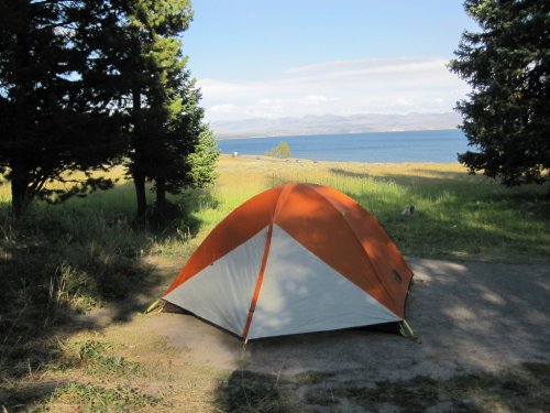 An orange and white tent set up near a lake, surrounded by trees and grassy areas under a clear blue sky.