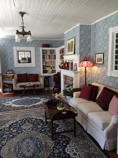 Cozy living room with floral wallpaper, a white sofa, bookshelves, and a lamp, featuring a patterned rug and decorative accents.