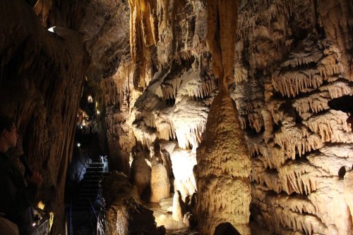 A dimly lit cave interior featuring stalactites and stalagmites, with a pathway and visitors in the background.