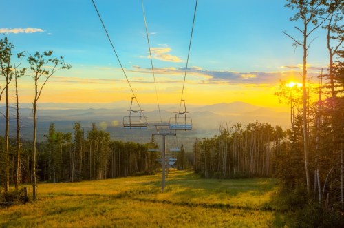 A scenic view of a ski lift at sunset, surrounded by trees and mountains under a colorful sky.