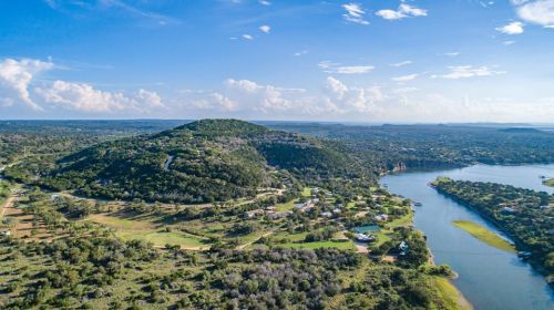 Aerial view of rolling hills and a winding river, surrounded by lush greenery and scattered homes under a blue sky.