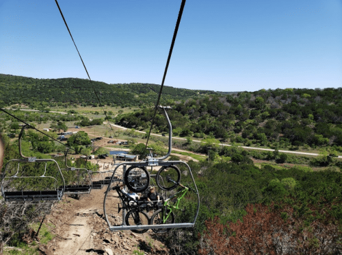 A chairlift with bicycles hanging below, overlooking a green landscape and a winding road in the distance.